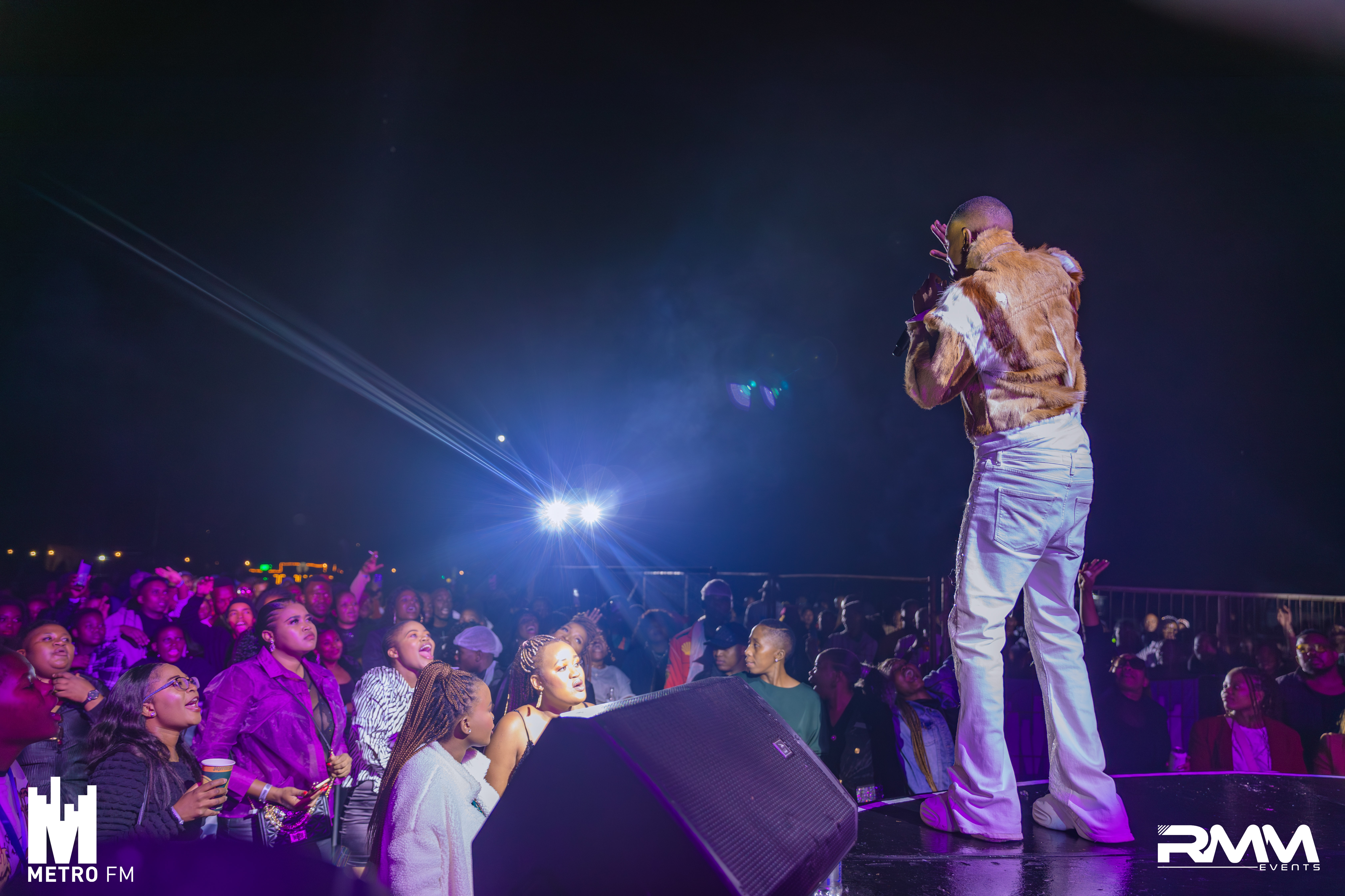 A person facing the crowd at a concert. White logos are on the  bottom corners of the image.