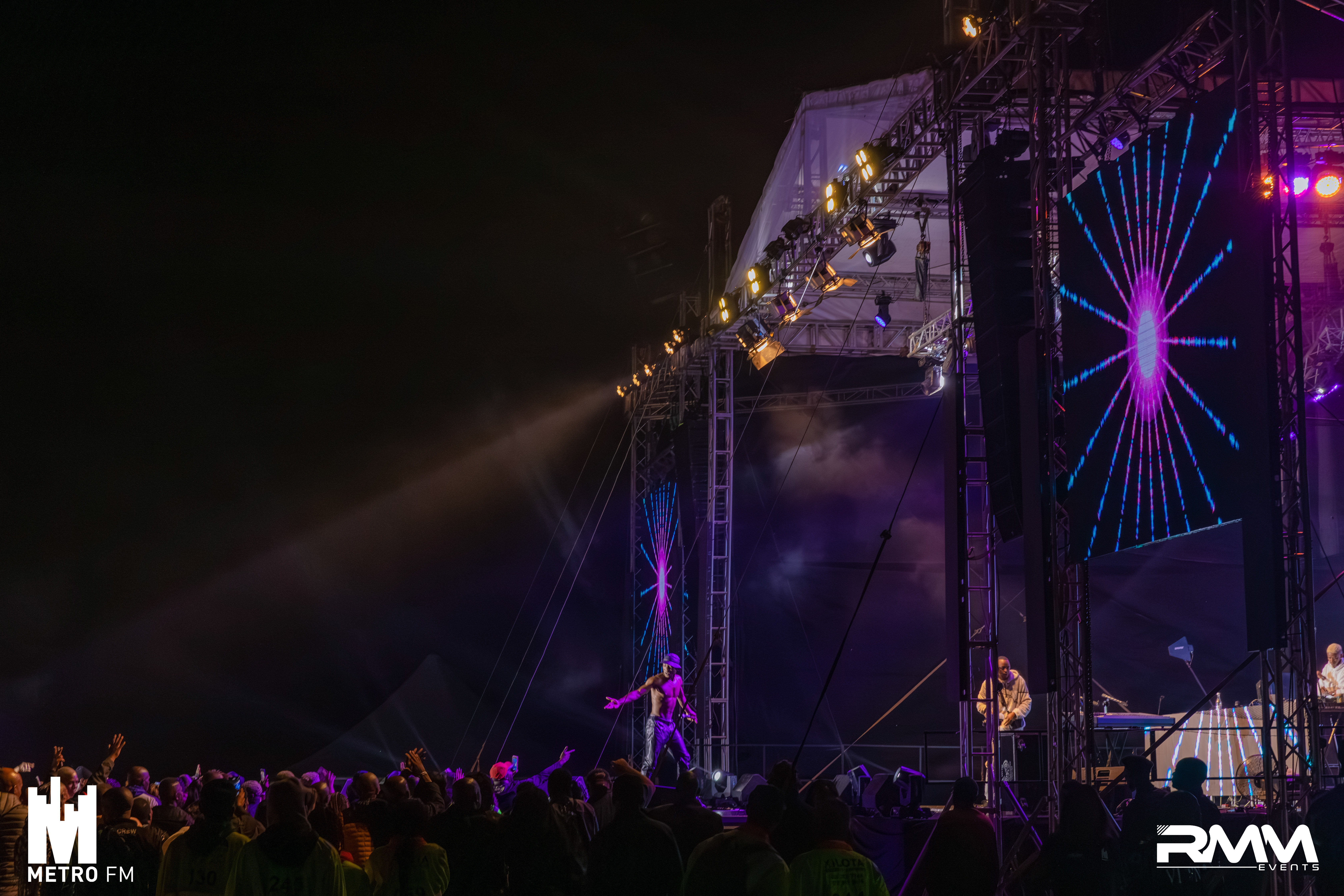 Concert with purple light show happening at night. White logos are on both bottom corners of image.